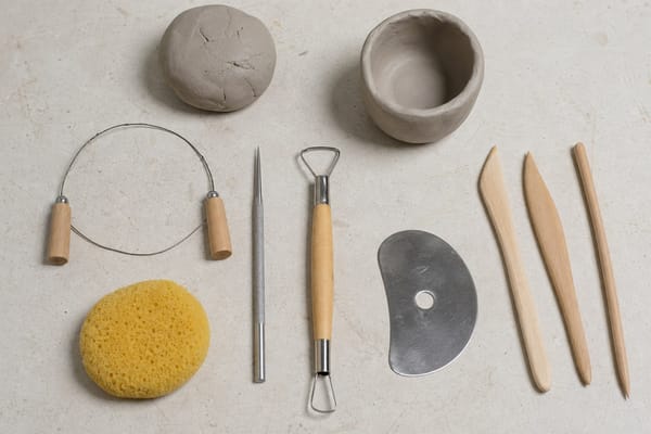 Beginner pottery tools including a wire clay cutter, needle tool, loop tool, pottery rib, sponge, and clay on a studio table.